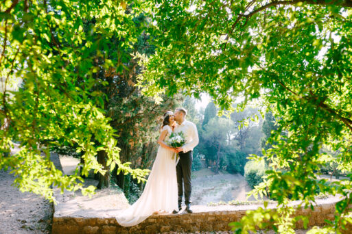 The bride and groom are hugging among beautiful lush branches of trees in the sunny day in the park. High quality photo
Свадьба летом