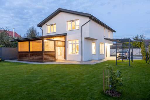 Exterior view of modern white house with courtyard, patio area, green grass lawn, garden and a car in the evening.
Загородный дом