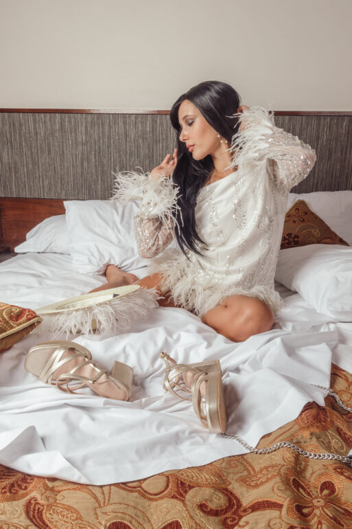 A young woman with brown hair sits on a hotel bed, wearing a long dress. The room is cozy with brown furnishings.
Богиня секс