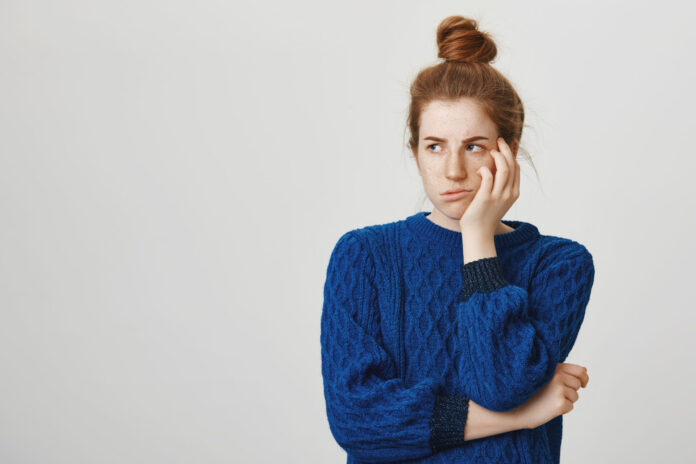 Jealous sister is offended that she did not got present. Gloomy annoyed redhead female student in winter sweater, turning head aside and leaning on palm, sulking and puckering being angry at someone